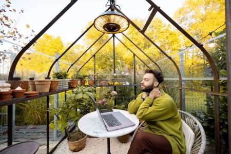 Photo for Man works on laptop while sitting by the round table in glasshouse with plants and flowers at backyard. Work from home at cozy atmosphere on nature - Royalty Free Image