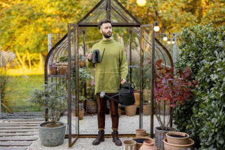 Foto de Retrato de un hombre elegante de pie con regadera frente a un pequeño invernadero para el cultivo de plantas en el jardín. Hobby, el trabajo en el concepto de jardín - Imagen libre de derechos