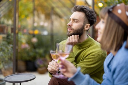 Téléchargez les photos : Jeune couple élégant traîner ensemble, parler et boire du vin tout en étant assis à l'arrière-cour. Homme et femme passer du temps en soirée à l'atmosphère confortable en plein air - en image libre de droit