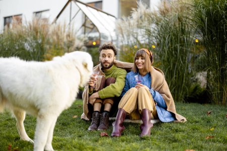 Photo for Young stylish couple warming up covered with plaid while sitting together and play with their cute dog on green lawn at backyard. Young family spending autumn time outdoors - Royalty Free Image