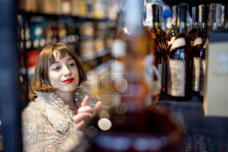 Téléchargez les photos : Woman chooses strong alcohol standing between rows of bottles in a supermarket. Buying cognac drink in alcohol shop - en image libre de droit