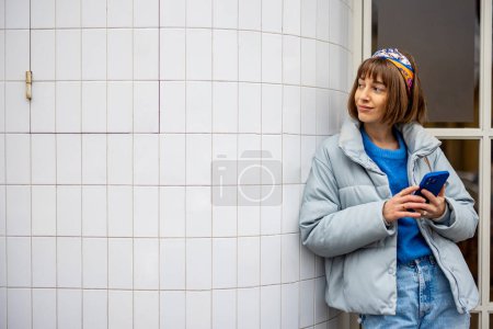 Foto de Mujer joven y elegante en azul ropa casual se para con el teléfono en el fondo de la pared de baldosas blancas al aire libre. Concepto de estilo de vida y estilo de ciudad - Imagen libre de derechos
