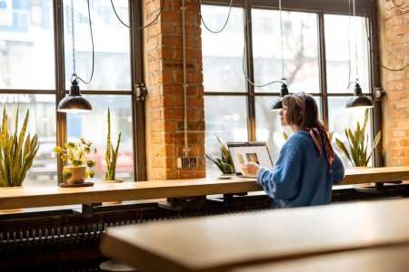 Photo for Young stylish woman works on laptop while sitting by the big window at modern coffee shop. Wide view from the backside - Royalty Free Image