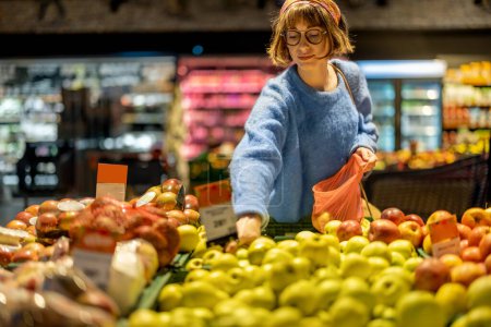 Téléchargez les photos : Young woman chooses apples to buy and puts them into mesh bag at fruit department of a supermarket. Concept of consumerism of healthy food and use of reusable bags - en image libre de droit