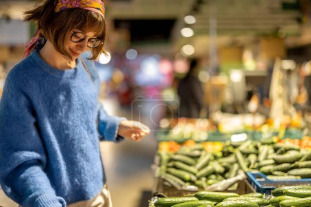 Foto de Young woman chooses cucumbers, buying vegetables at supermarket. Concept of consumerism and healthy lifestyle - Imagen libre de derechos