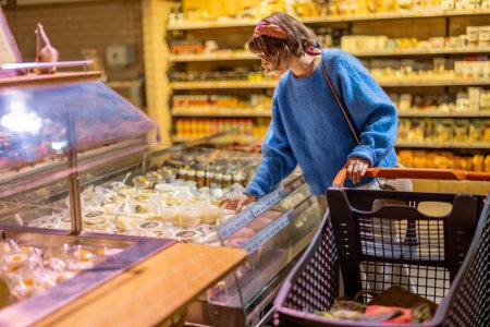 Foto de Young woman buying cheese, standing with shopping trolley in supermarket. Concept of shopping dairy products in shop - Imagen libre de derechos