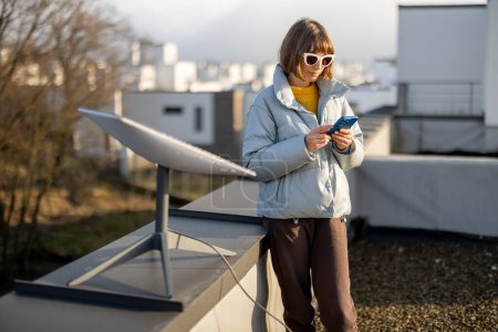 Téléchargez les photos : LVIV, UKRAINE - January, 2023: Woman with phone uses Starlink satellite Internet, constellation operated by SpaceX on the roof of her house with satellite dish in front - en image libre de droit