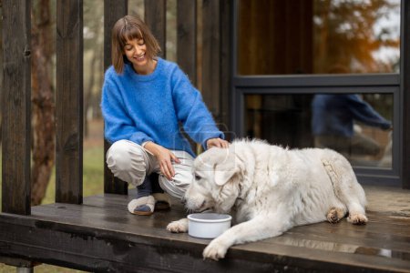 Téléchargez les photos : Young woman feeds her cute white dog while sitting together on a porch of wooden house in the forest. Pets caring and rest on nature concept - en image libre de droit