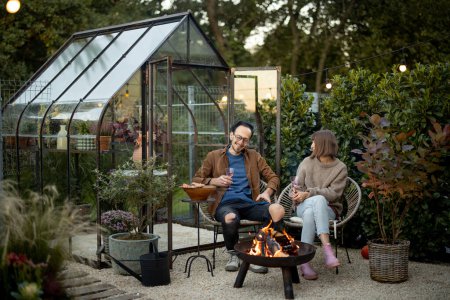 Photo for Young couple having fun during romantic and atmospheric dinner by the fire at backyard with vintage glasshouse behind - Royalty Free Image