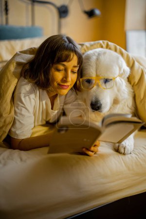 Photo for Young woman with her cute dog in eyeglasses reading a book while lying togther under a blanket in bedroom. Spending leisure time, friendship with pet concept - Royalty Free Image