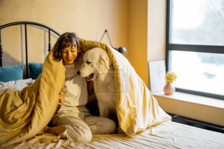 Foto de Cute young woman playing with her white adorable dog while sitting together covered with blanket on bed at home. Concept of friendship with pets and home coziness - Imagen libre de derechos