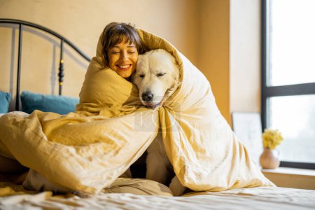 Foto de Cute young woman hugs with her white adorable dog, covered together with blanket on bed at home. Concept of friendship with pets and home coziness - Imagen libre de derechos
