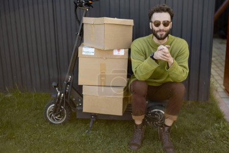 Foto de Retrato de un hombre sentado en scooter eléctrico con cajas de cartón para entregar al aire libre. Concepto de entrega y estilo de vida sostenible - Imagen libre de derechos