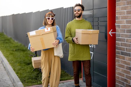 Foto de Alegre joven hombre y mujer llevan paquetes de cartón juntos recibidos en la máquina de correo automático al aire libre. Concepto de tecnologías modernas de entrega - Imagen libre de derechos