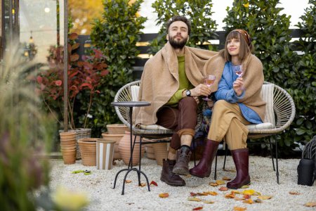 Téléchargez les photos : Young stylish couple warming up together, sitting covered with plaid by the fire and drink wine, spending evening time at cozy atmosphere in beautiful garden - en image libre de droit