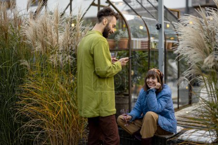 Téléchargez les photos : Young couple hang out, uising phone and drink wine on a porch with lush pampas grass on backyard of their house. Couple spend autumn time outdoors - en image libre de droit