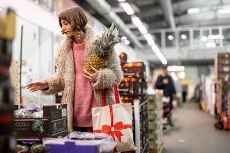 Foto de Woman buying fruits at local indoor market choosing ananas during winter time. Girl in fur coat shopping fruits for the holidays - Imagen libre de derechos