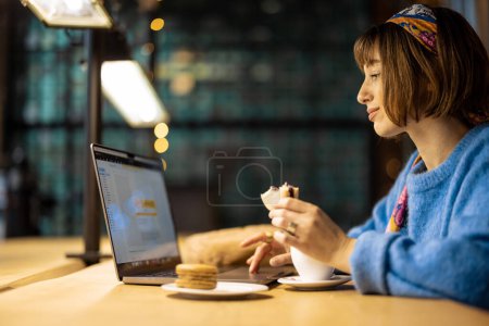 Foto de Young stylish woman works on laptop while sitting with a coffee drink at modern cafe. Concept of remote work from public place, digital freelance and modern lifestyle - Imagen libre de derechos