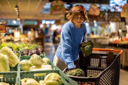 Téléchargez les photos : Young woman chooses broccoli, buying vegetables in supermarket. Concept of shopping groceries and healthy lifestyle - en image libre de droit
