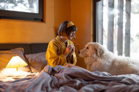 Téléchargez les photos : Young woman cares her huge adorable white dog while lying in bed in tiny bedroom of wooden cabin on nature. House coziness and friendship with pets concept - en image libre de droit