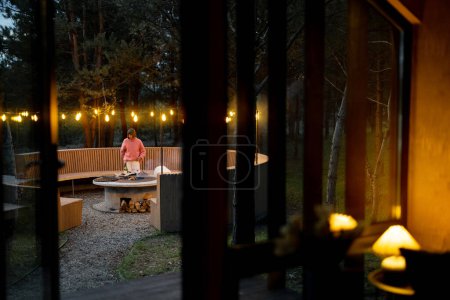 Photo for Beautiful lounge area with round bench and bonfire illuminated with garlands near house in pine forest, view from the house - Royalty Free Image