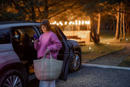 Téléchargez les photos : Woman arrives by car to a house in forest, standing with bag and phone near vehicle in the evening time. Traveling by car and rest in cabins on nature concept - en image libre de droit