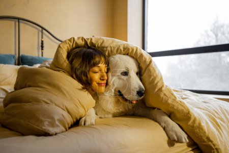 Téléchargez les photos : Jeune femme étreint avec son chien blanc adorable tout en étant couché couvert de couverture dans le lit à la maison. Concept d'amitié avec les animaux domestiques et de confort à la maison - en image libre de droit