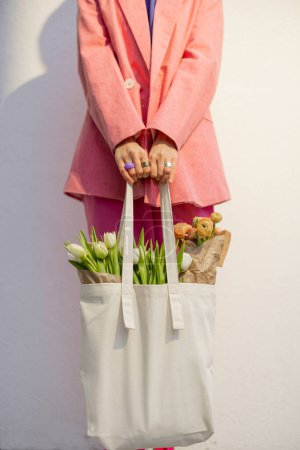 Foto de Stylish woman in pink clothes with eco handbag full of flowers on white background, close-up, with no face. Canvas bag with blank space - Imagen libre de derechos