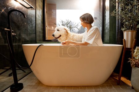 Foto de Mujer joven lavando su lindo perro blanco en la bañera en casa. Concepto de cuidado de animales, procedimientos de spa para mascotas y amistad - Imagen libre de derechos