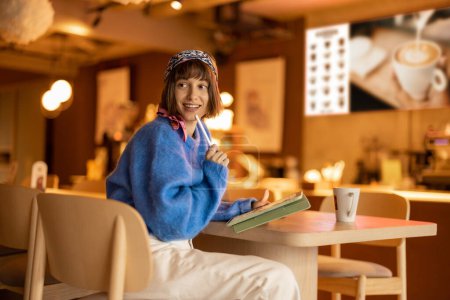 Téléchargez les photos : Young adult woman works on a digital tablet while sitting at modern coffee shop. Concept of remote creative work online - en image libre de droit
