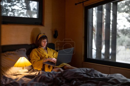Photo for Woman works on laptop while lying in bed late in the evening at tiny bedroom of wooden cabin on nature. Online work from cozy house during bedtime - Royalty Free Image