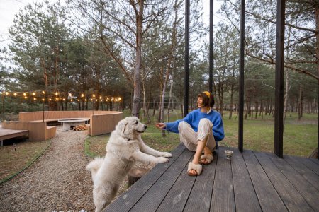 Foto de Young woman cares her dog while sitting on a porch of wooden house in pine forest, spending leisure time together and resting at countryside - Imagen libre de derechos