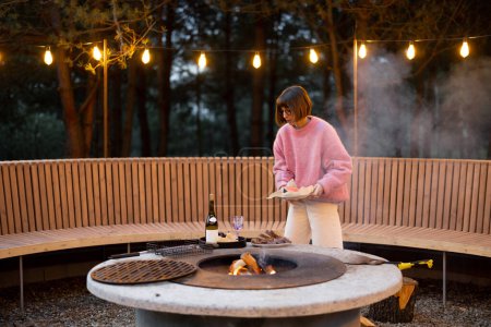 Photo for Young woman prepares food on beautiful outdoor lounge area with fire and round bench in pine forest at dusk. Luxury lifestyle at countryside concept - Royalty Free Image