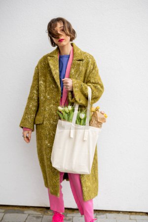 Foto de Portrait of a stylish woman in bright clothes with eco handbag full of flowers on white background. Canvas bag with blank space - Imagen libre de derechos