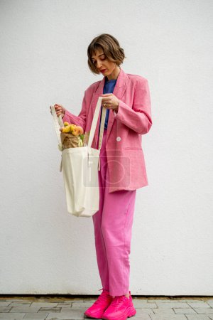 Foto de Stylish woman in pink suit with eco handbag and flowers on white background. Canvas bag with blank space - Imagen libre de derechos