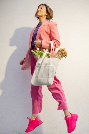 Foto de Stylish woman in pink suit jumps with eco handbag full of flowers on white background. Canvas bag with blank space. Concept of style and spring time - Imagen libre de derechos