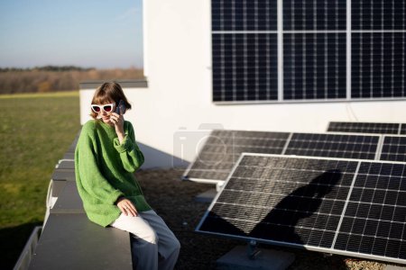 Foto de Mujer habla por teléfono mientras está sentada en el techo de su casa con una estación solar instalada en ella. Feliz propietario de un hogar independiente de la energía - Imagen libre de derechos