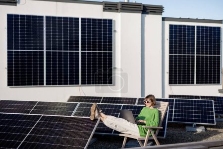 Téléchargez les photos : Woman works on laptop and phone while sitting on rooftop with a solar station. Concept of remote work, alternative energy and sustainable lifestyle - en image libre de droit