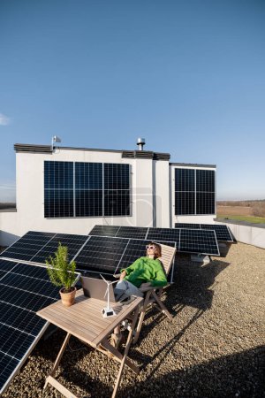 Foto de Woman works on laptop while sitting relaxed by the table on rooftop with a solar power plant, wide view. Concept of remote work, alternative energy and sustainable lifestyle - Imagen libre de derechos