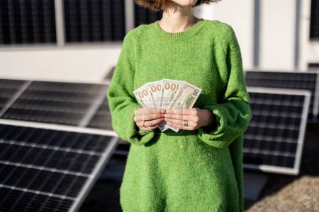 Foto de La mujer cuenta los billetes de dólar ahorrados debido a la generación de energía de una planta de energía solar instalada en la azotea de su casa. Concepto de inversión en energías alternativas - Imagen libre de derechos