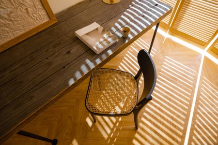Foto de Vista desde arriba sobre mesa de madera y silla en el suelo hecha de parquet con espina de arenque. Sombras de las persianas de las ventanas. Fragmento del interior en tonos beige en estilo boho - Imagen libre de derechos