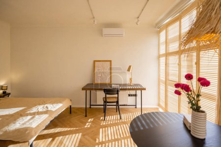 Foto de Elegante y soleado apartamento estudio en tonos beige con mesa de comedor, lugar de trabajo y cama. Hogar acogedor y concepto de diseño de interiores. Foto real - Imagen libre de derechos