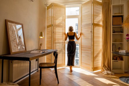 Téléchargez les photos : Woman opens window blinds letting the sun inside the room, spending good morning in sunny and cozy apartment in beige tones. Interior view - en image libre de droit