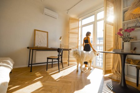 Photo for Young woman stands with her dog by the window blinds and looks away in cozy and sunny living room of modern apartment in beige tones. Quarantine, loneliness and life at home concept - Royalty Free Image