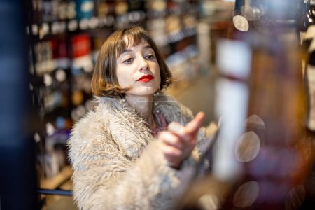Foto de Woman chooses strong alcohol standing between rows of bottles in a supermarket. Buying cognac drink in alcohol shop - Imagen libre de derechos