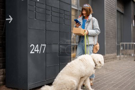 Photo for Young woman uses phone while receiving a parcel from automatic post office machine during a walk with her dog in city. Concept of modern technologies in delivery services and lifestyle - Royalty Free Image