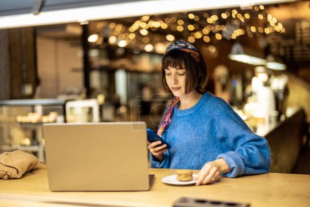 Foto de Young stylish woman works on laptop while sitting with a coffee drink at modern cafe. Concept of remote work from public place, digital freelance and modern lifestyle - Imagen libre de derechos