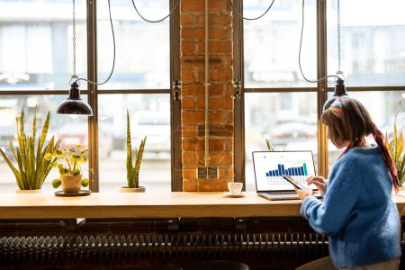 Photo for Young stylish woman works on laptop and phone while sitting by the big window at modern coffee shop. Wide view from the backside - Royalty Free Image