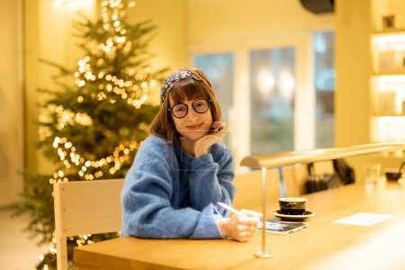 Téléchargez les photos : Portrait de jeune femme adulte assise au café moderne dans des tons jaunes et arbre de Noël sur fond. Concept de travail créatif à distance en ligne - en image libre de droit