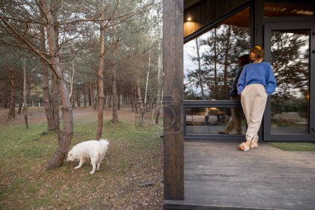 Téléchargez les photos : Young woman stands on porch of a wooden house in pine forest, enjoying nature, resting with a dog in cottage at countryside - en image libre de droit